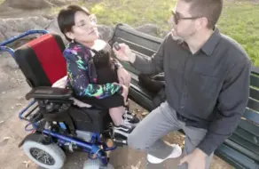 Antonio Mallorca con gafas y camisa gris entrevistando a una chica en silla de ruedas sentada en un banco del parque